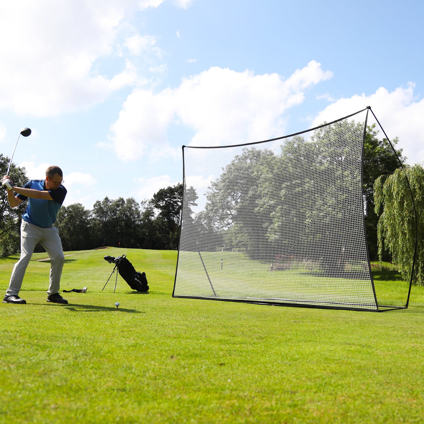 Golfer in a blue shirt starting his backswing on a teed ball in front of the MEGA NET Golf Hitting Net 12x9' on a fairway.