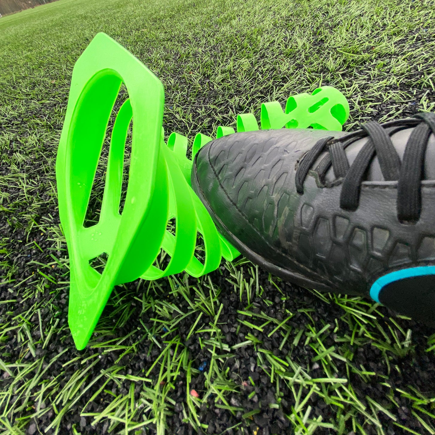 Close-up of a football boot pressing a green 9" collapsible cone flat on artificial grass.