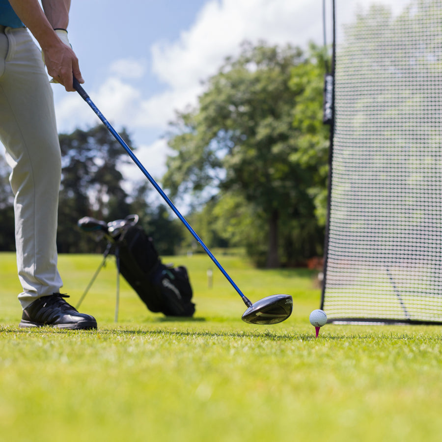 Golfer lining up a driver behind a teed ball on grass, next to the edge of the MEGA NET Golf Hitting Net 12x9'.