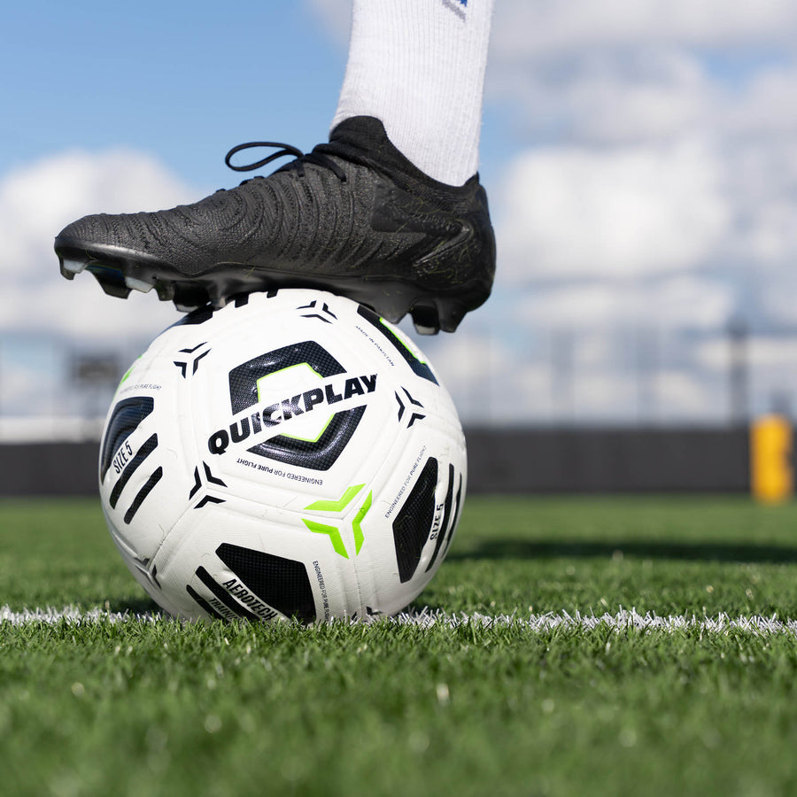 Football with 'QuickPlay' branding on a grass field, foot wearing a black boot on top.