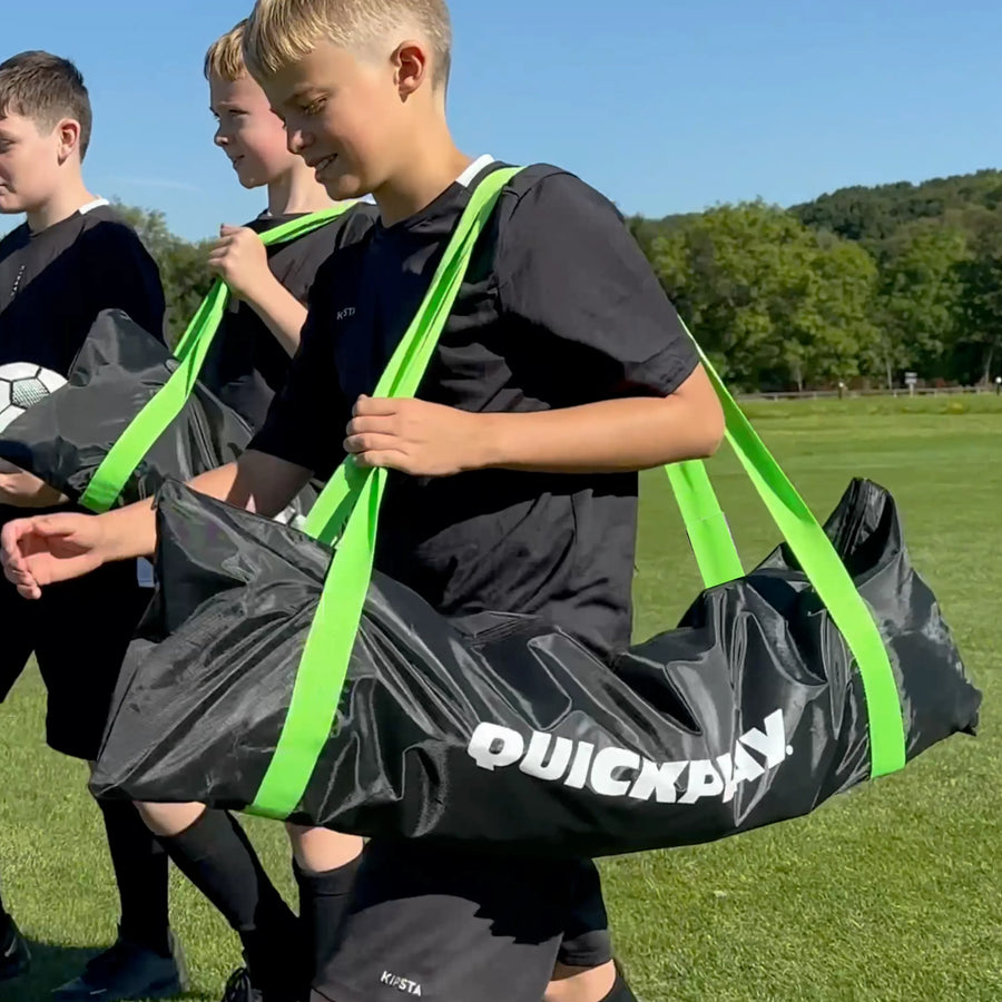 Boys carrying black QUICKPLAY Equipment & Goal Carry Bags (LG) with green handles across a grass field.