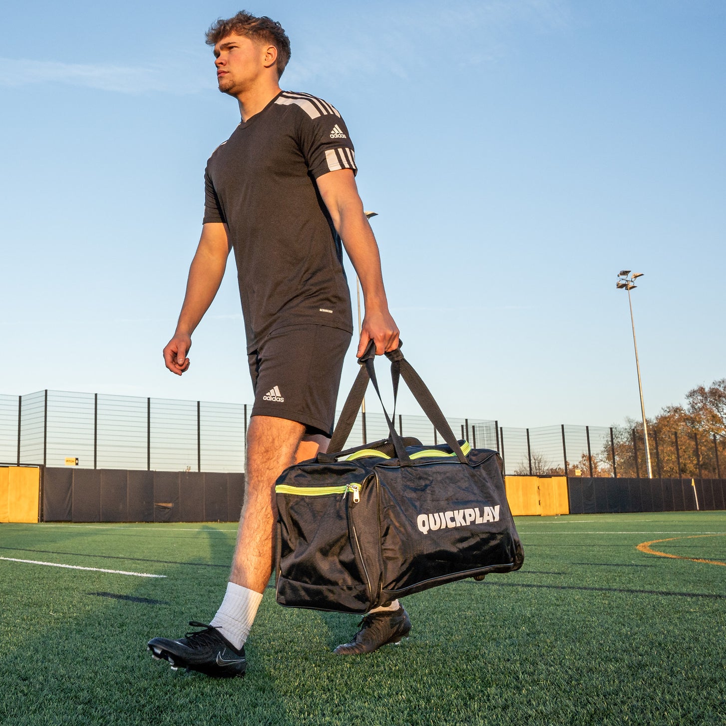Player in kit walking across an artificial pitch carrying the QUICKPLAY Equipment & Kit Bag (M) by its handles.