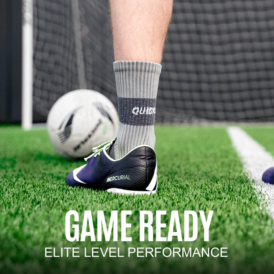 Player's foot in a grey QUICKPLAY Grip Sock and a football boot beside a ball on artificial grass, with the text "Game ready".