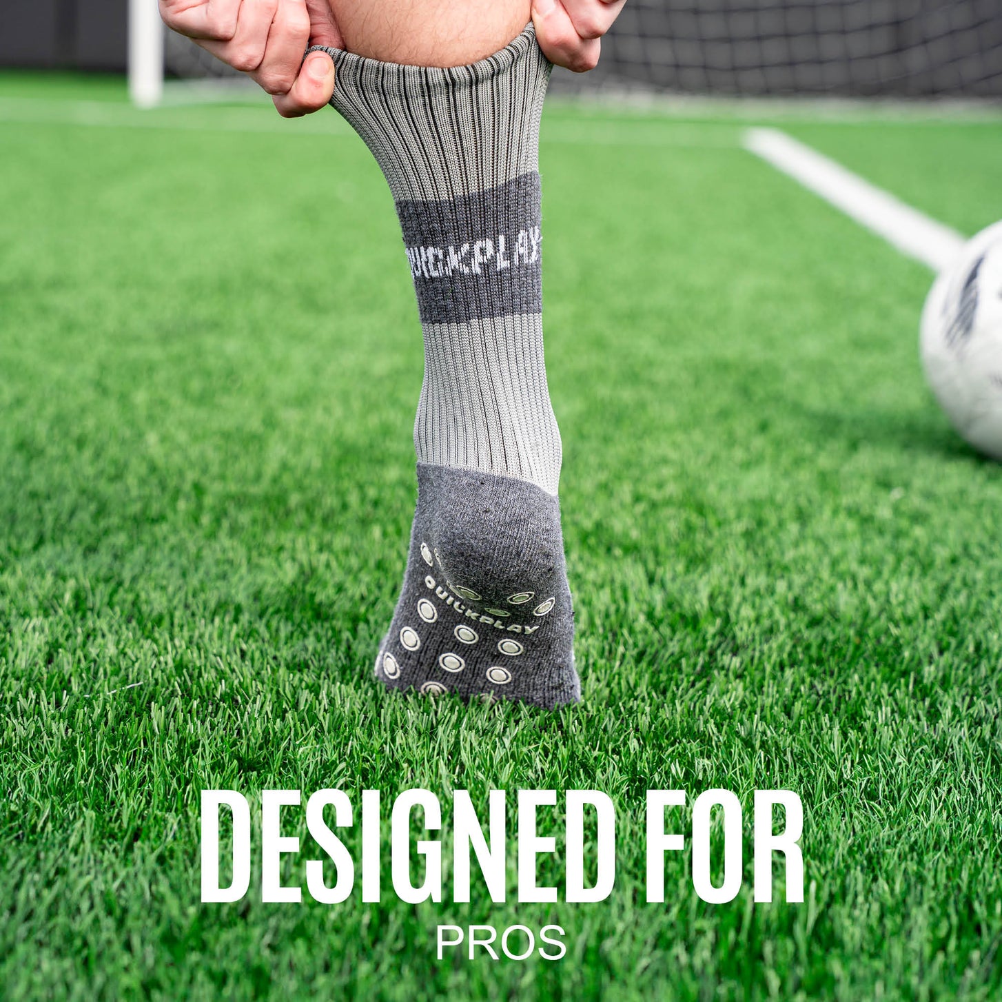 Player pulling a grey QUICKPLAY Grip Sock up their leg on artificial grass beside a ball, with the text "Designed for pros".