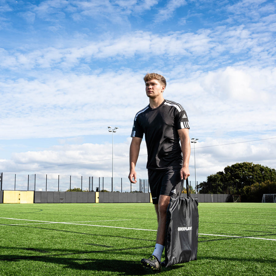 Player walking across an artificial pitch carrying a black QUICKPLAY bag for the Handheld Rebounder.