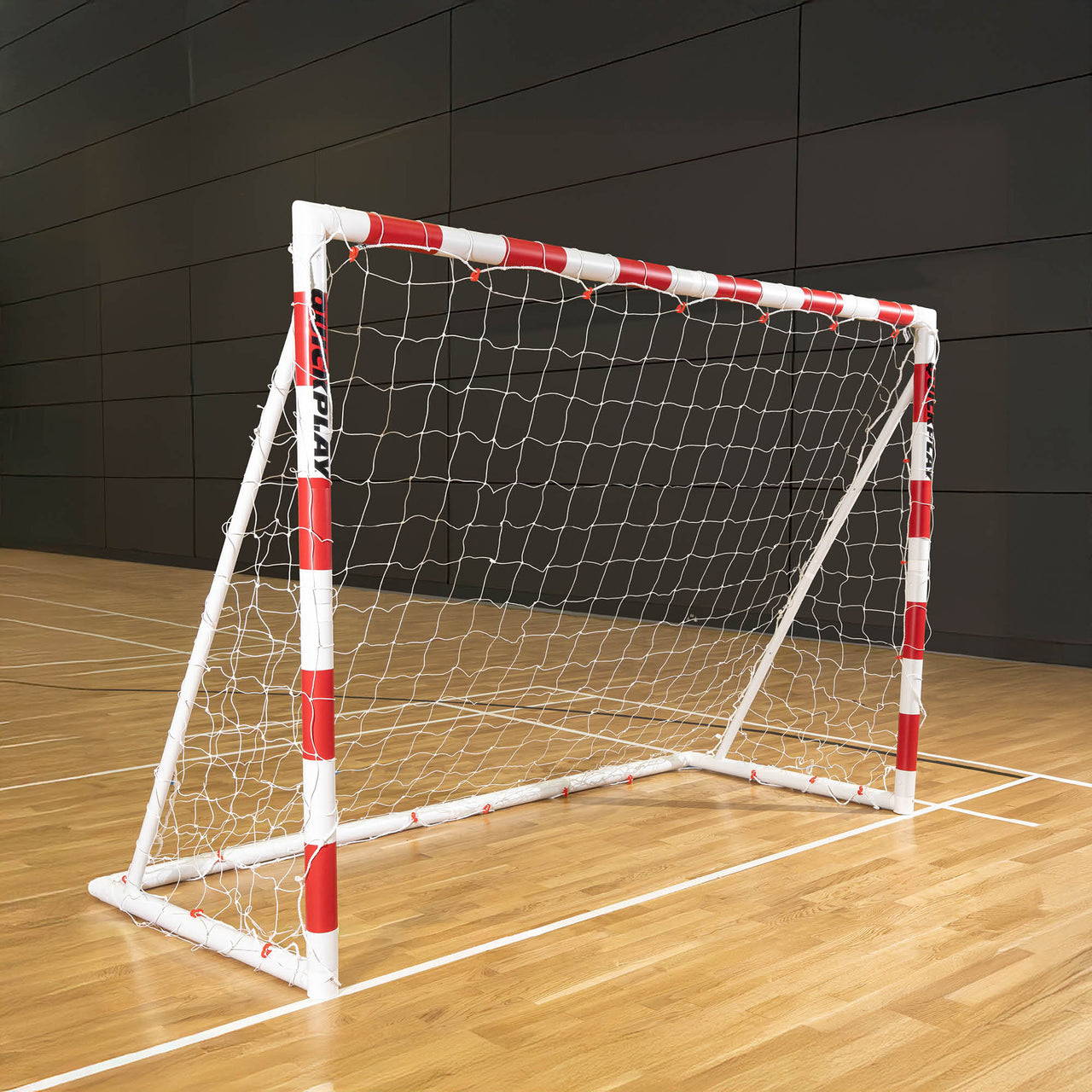 Small handball goal with red and white frame on a wooden floor against a dark panelled wall.