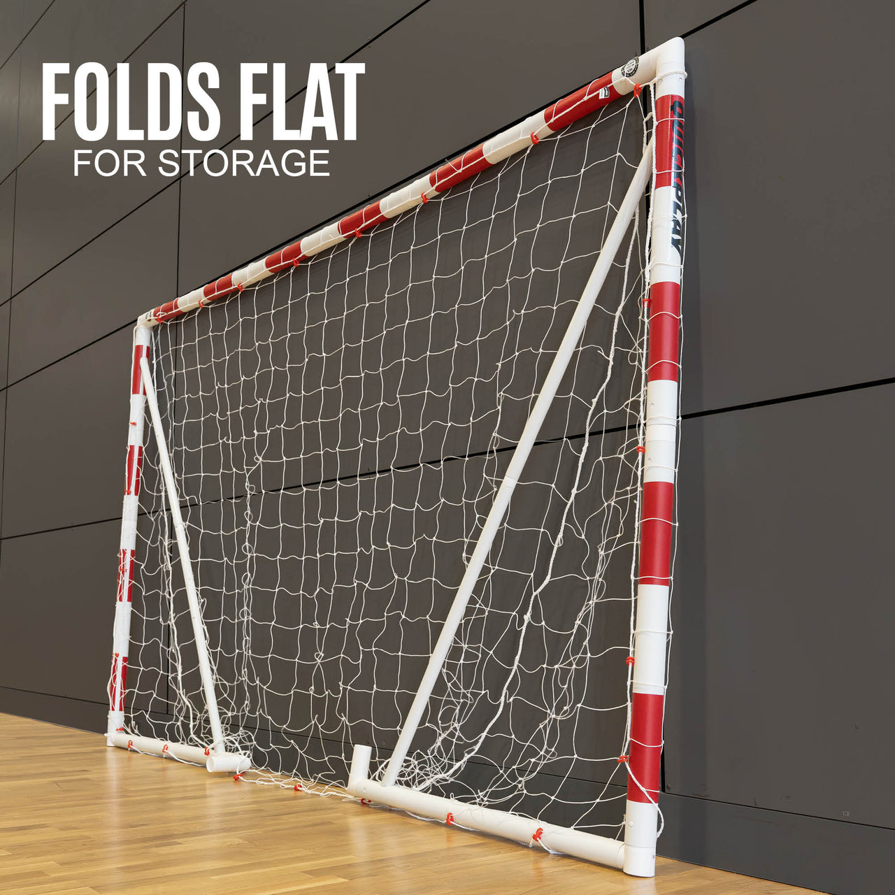 Portable handball goal with red and white design on a wooden floor against a dark panelled wall.
