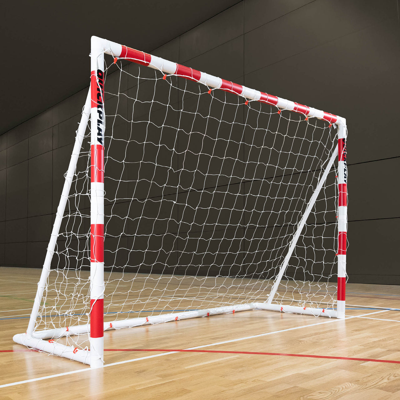 Small handball goal with red and white frame on a wooden floor against a dark wall.