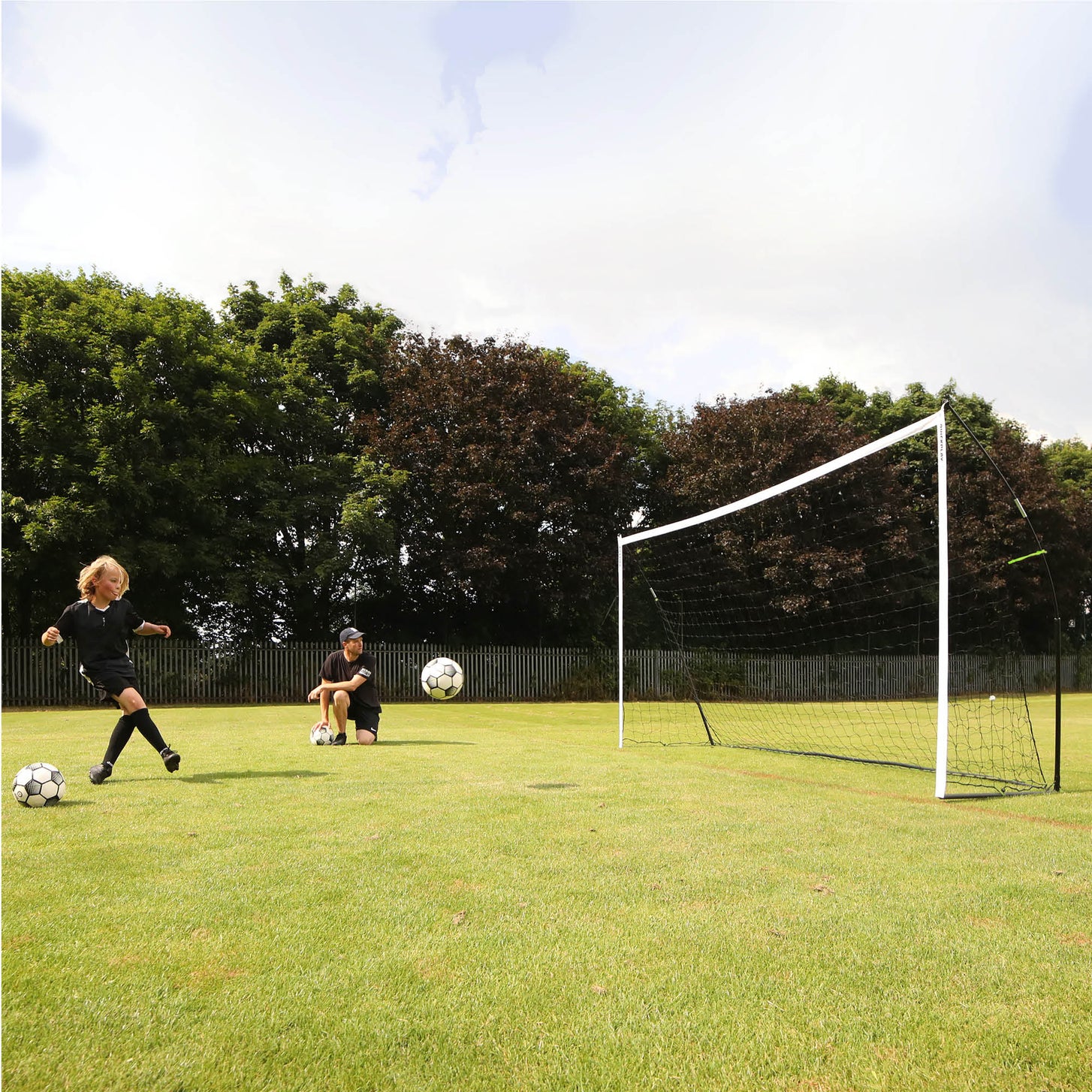 KICKSTER goal on grass as a girl kicks a ball and a man kneels nearby with another ball.