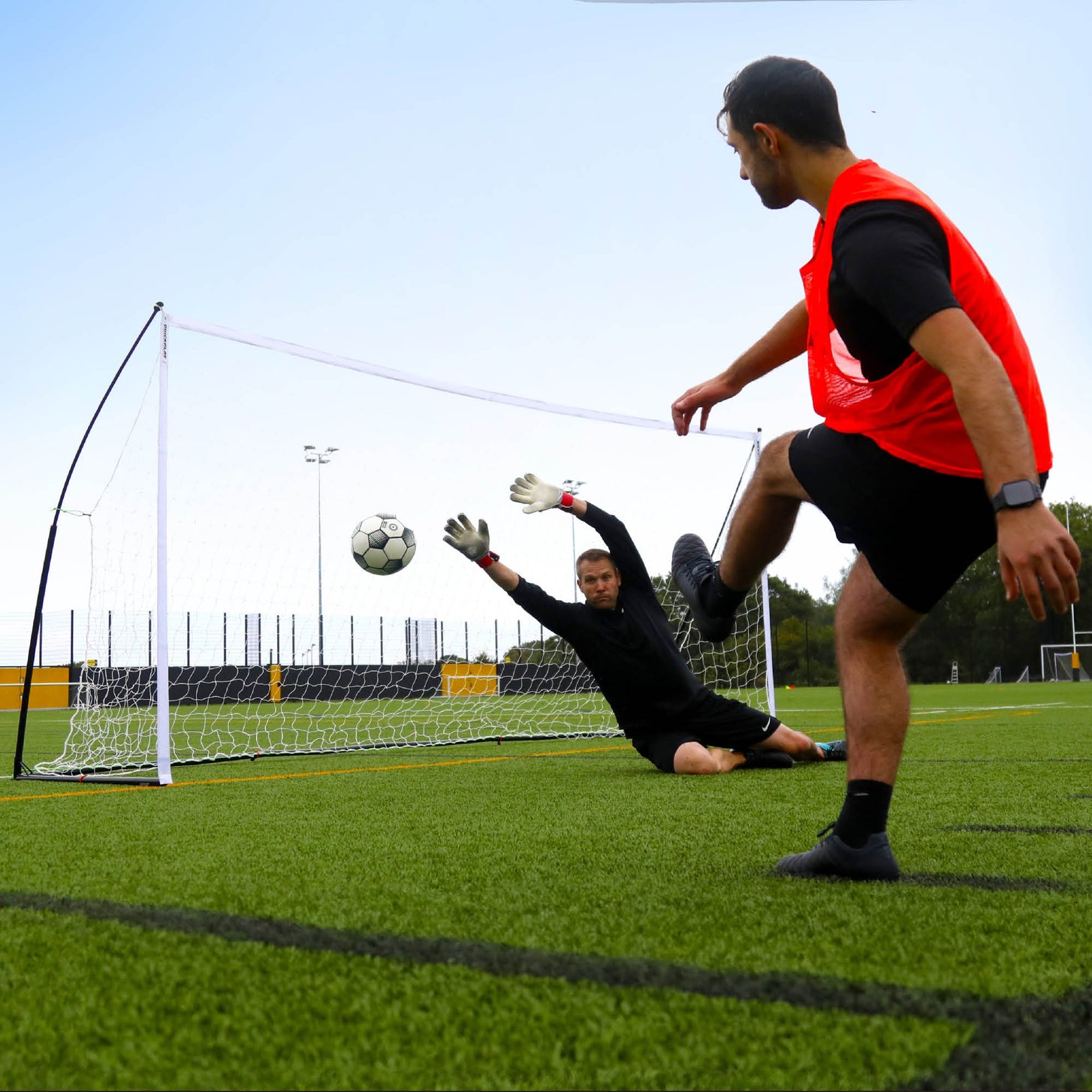 Player in an orange bib shooting past a diving goalkeeper into the KICKSTER Elite 12x6' goal on an artificial pitch.