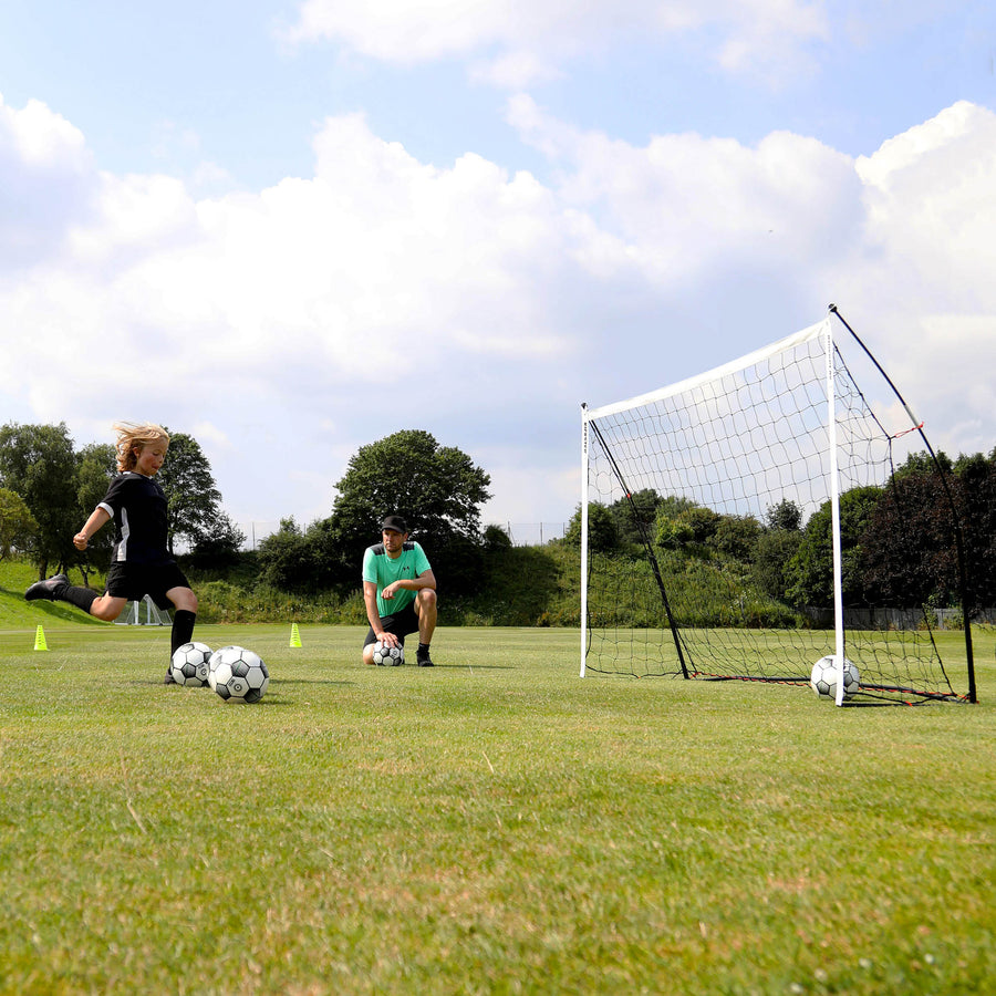 Child shooting at the KICKSTER 8x5' goal on a grass field while a man in a green top kneels and watches.