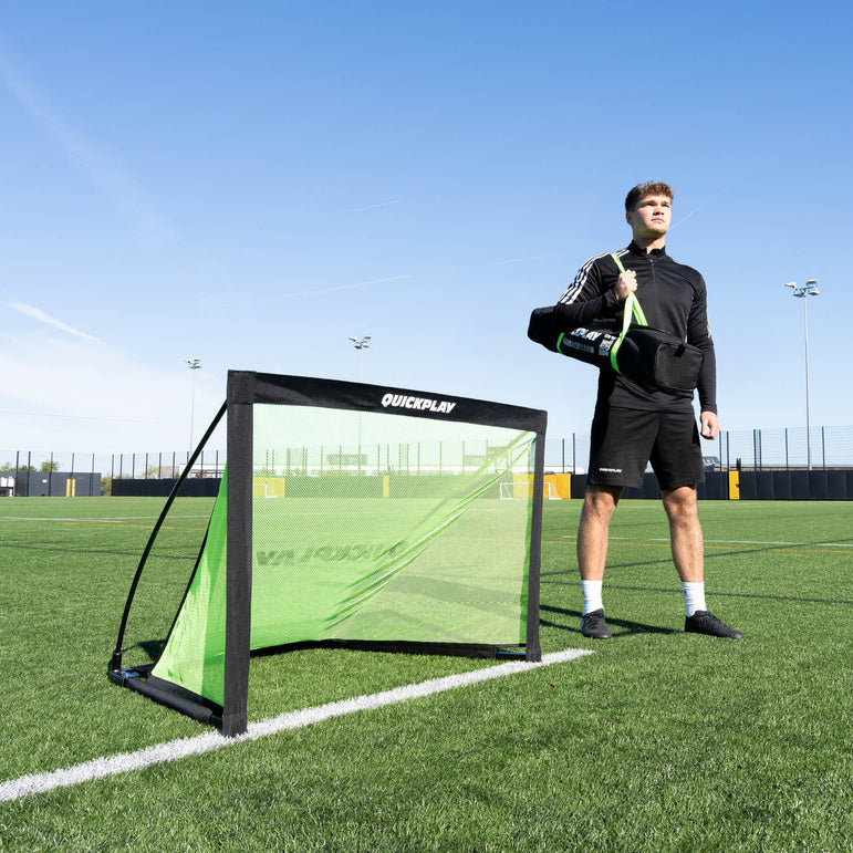 Person standing next to a green football goal on a pitch with clear blue sky.