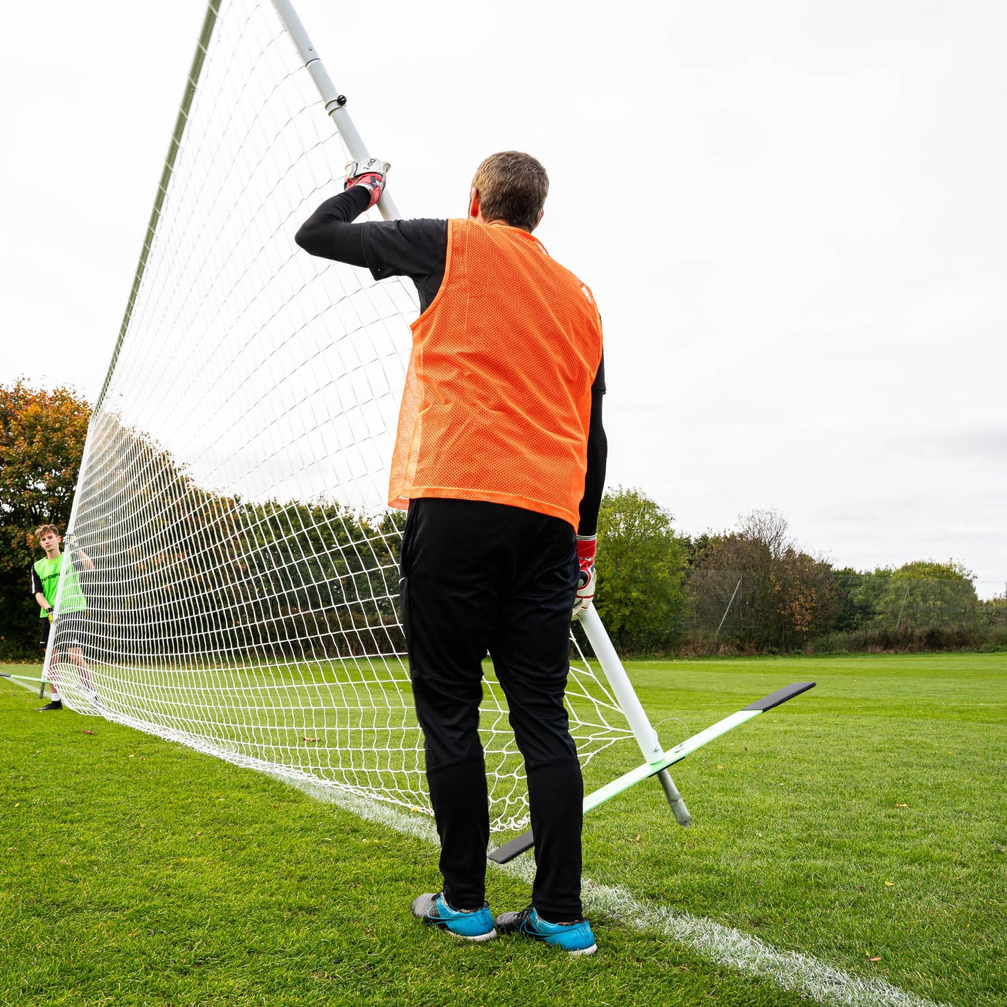 Coach in an orange bib holding the post of the PRO ALU Training Frame 18.5x6.5' on a grass pitch while a player watches.