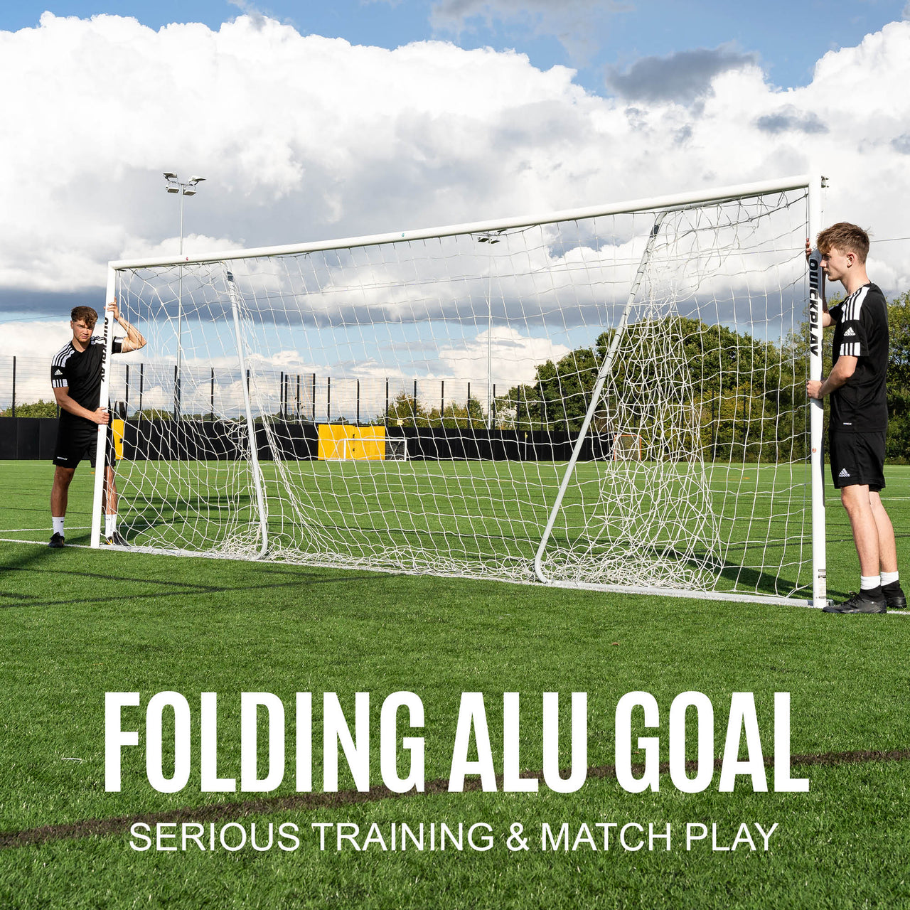 Two people setting up a folding aluminum goal on a grass field with a cloudy sky.