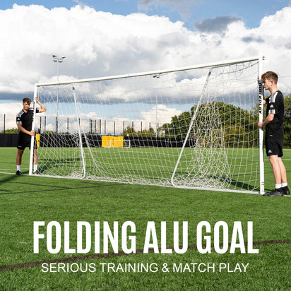 Two people setting up a folding aluminum goal on a grass field with a cloudy sky.
