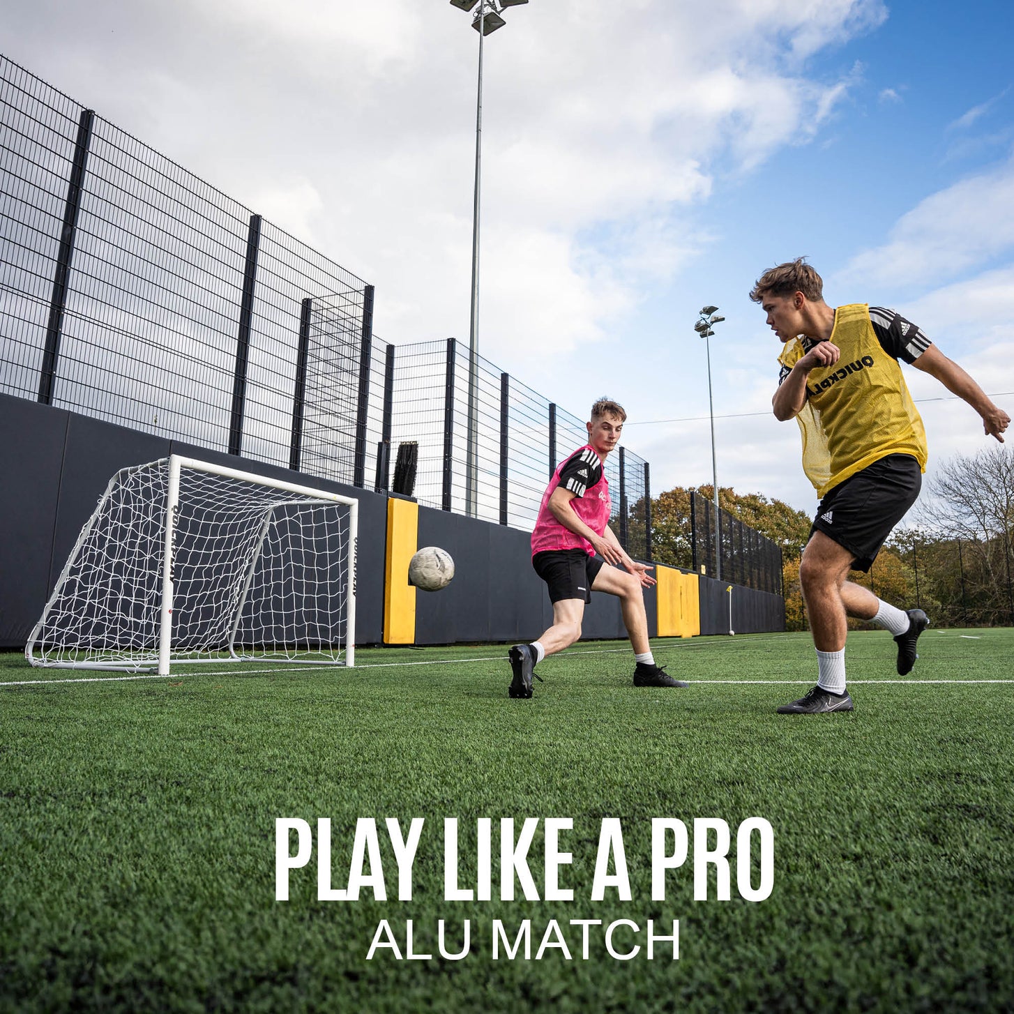 Two players in bibs passing in front of the PRO ALU Match60 6x4' goal on a fenced pitch, with the text "Play like a pro, ALU Match".