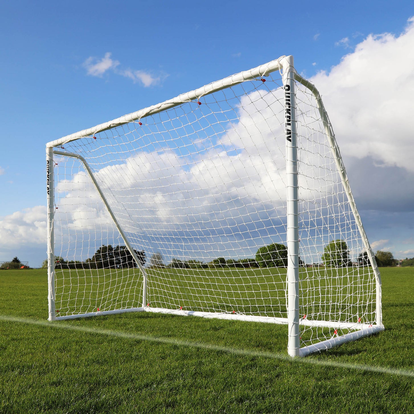 Q-FOLD Match 3x2m futsal goal with a white frame and net standing on a grass pitch under a blue sky, seen from the side.