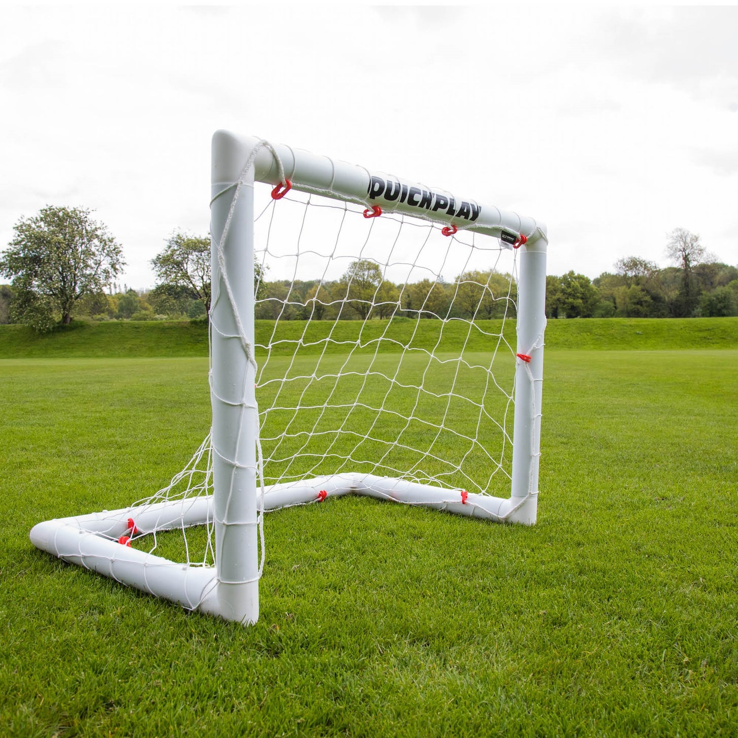 Q-FOLD Mini 3x2.5' goal seen at an angle on a grass field, showing the white frame, net and red net clips.