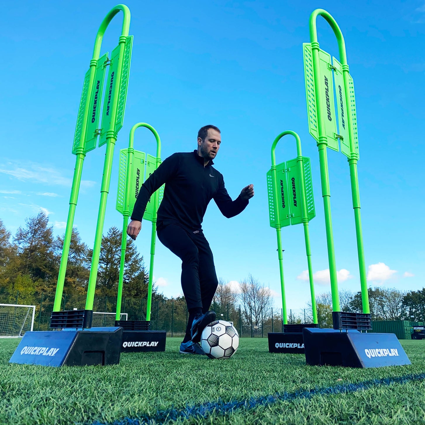 Player dribbling a ball through four REPLAY Defender mannequins on black bases, low angle on an artificial pitch.