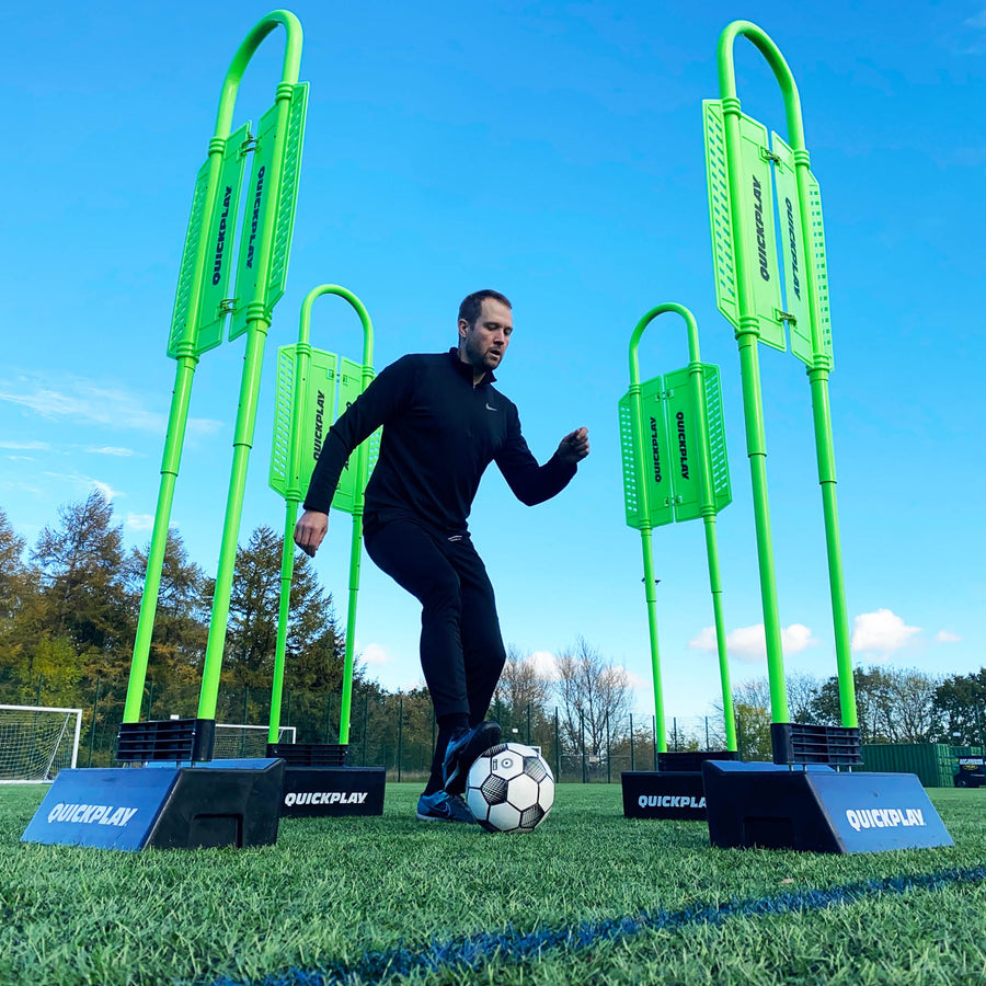 Player dribbling a ball through four REPLAY Defender mannequins on black bases, low angle on an artificial pitch.