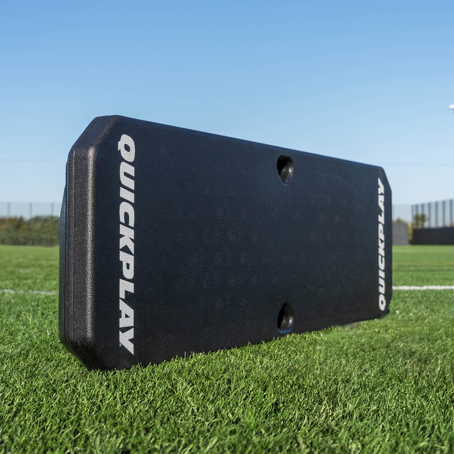 Black QuickPlay sports training rebounder on a grass field with a clear blue sky.