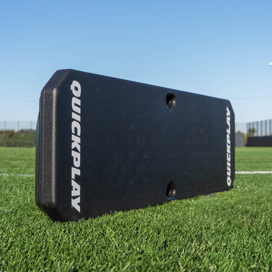 Black QuickPlay sports training rebounder on a grass field with a clear blue sky.