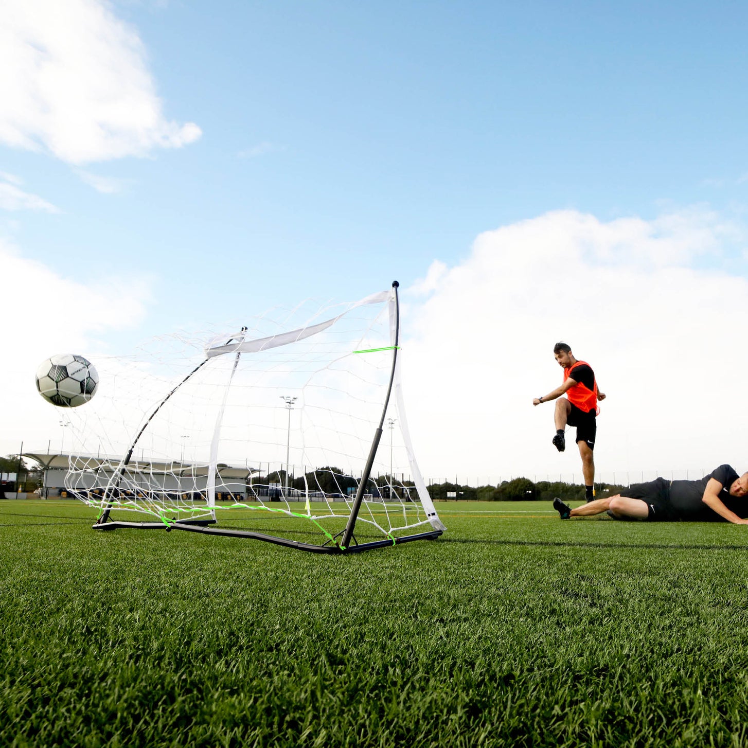 Player in an orange bib shooting on an artificial pitch, ball in the air beside a small white portable goal, another player on the ground.