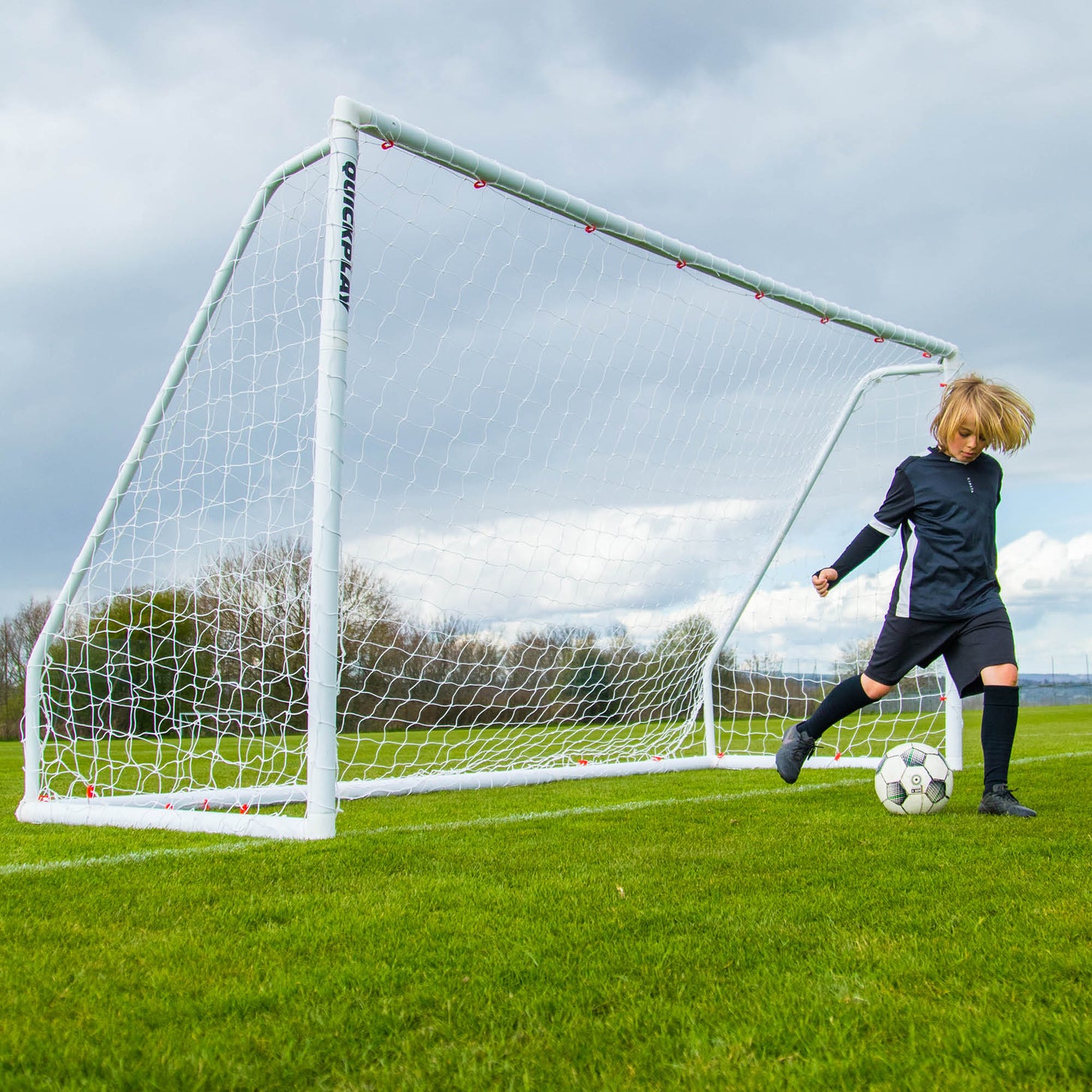 Child dribbling a ball beside the Q-FOLD Match 16x7' goal on a grass field under a cloudy sky.