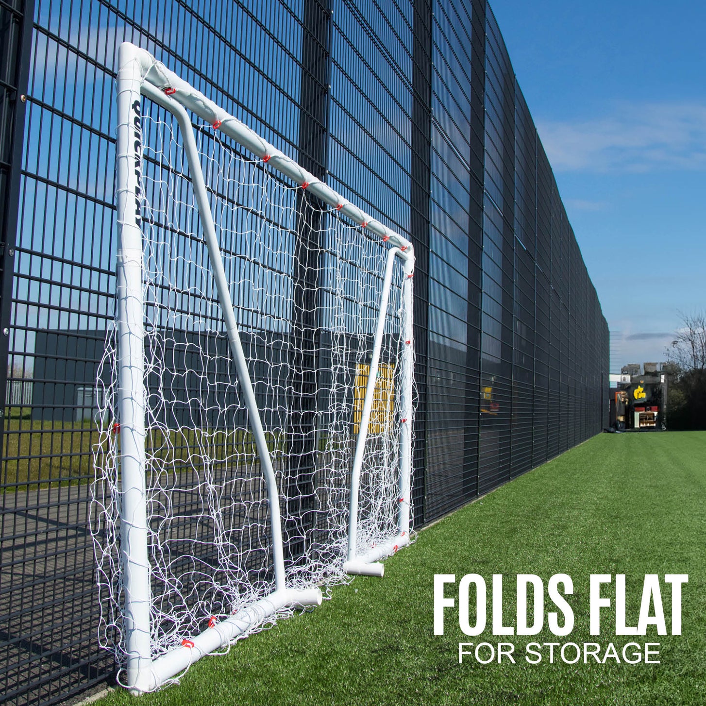 Q-FOLD Match 3x2m goal folded flat against a tall black fence beside an artificial pitch, with the text "Folds flat for storage".