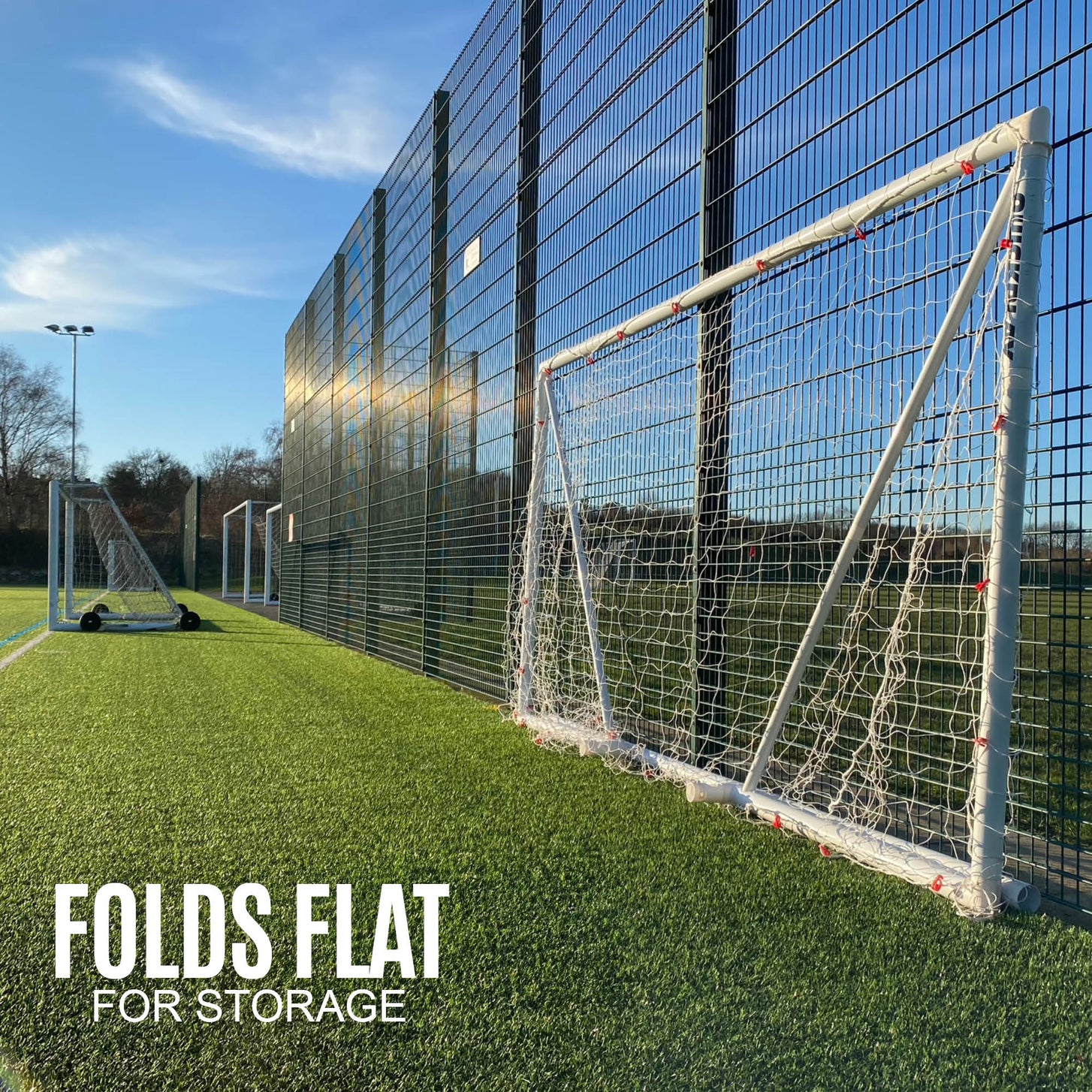 Q-FOLD Match 8x5' goal folded flat against a fence on an artificial pitch, with the text "Folds flat for storage".
