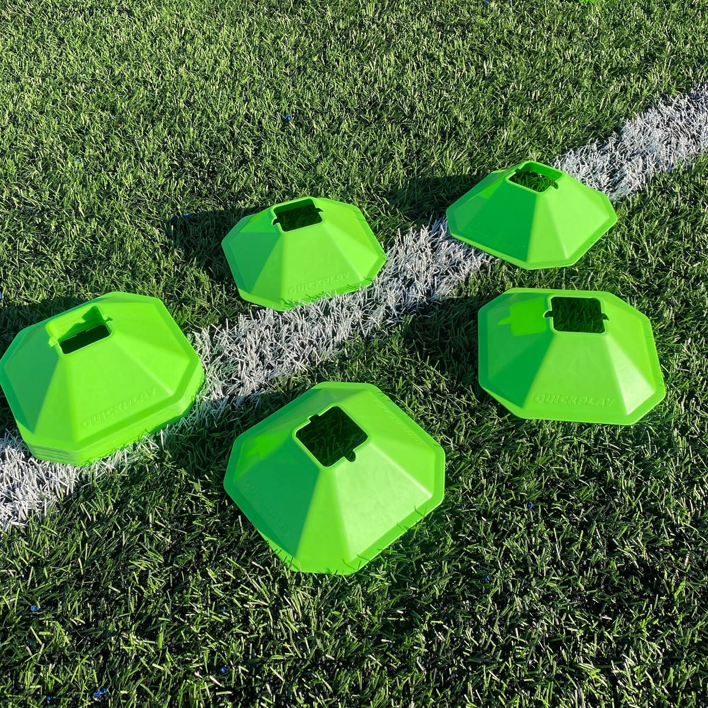 Five green Standard Cones spread out on an artificial pitch across a white line.