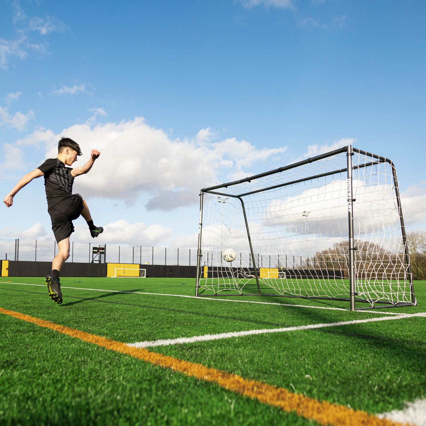 Young player shooting at the GS Series Football Goal 6x4' on an artificial pitch, with the ball hitting the net.