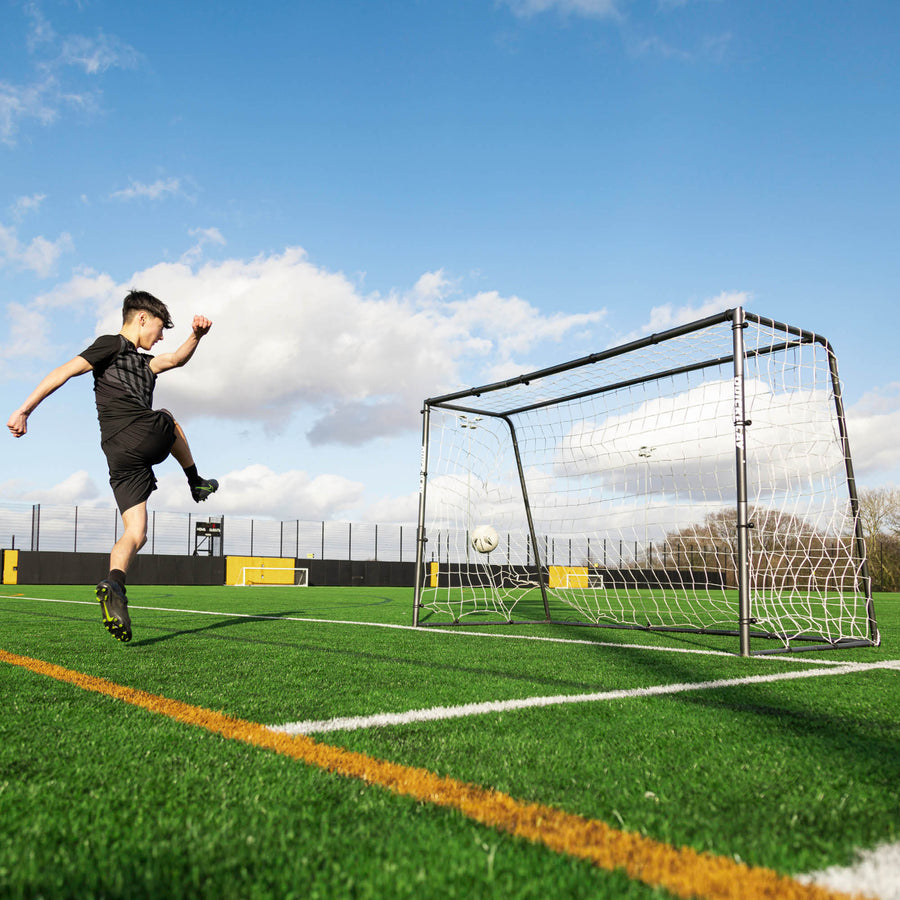 Young player shooting at the GS Series Football Goal 6x4' on an artificial pitch, with the ball hitting the net.