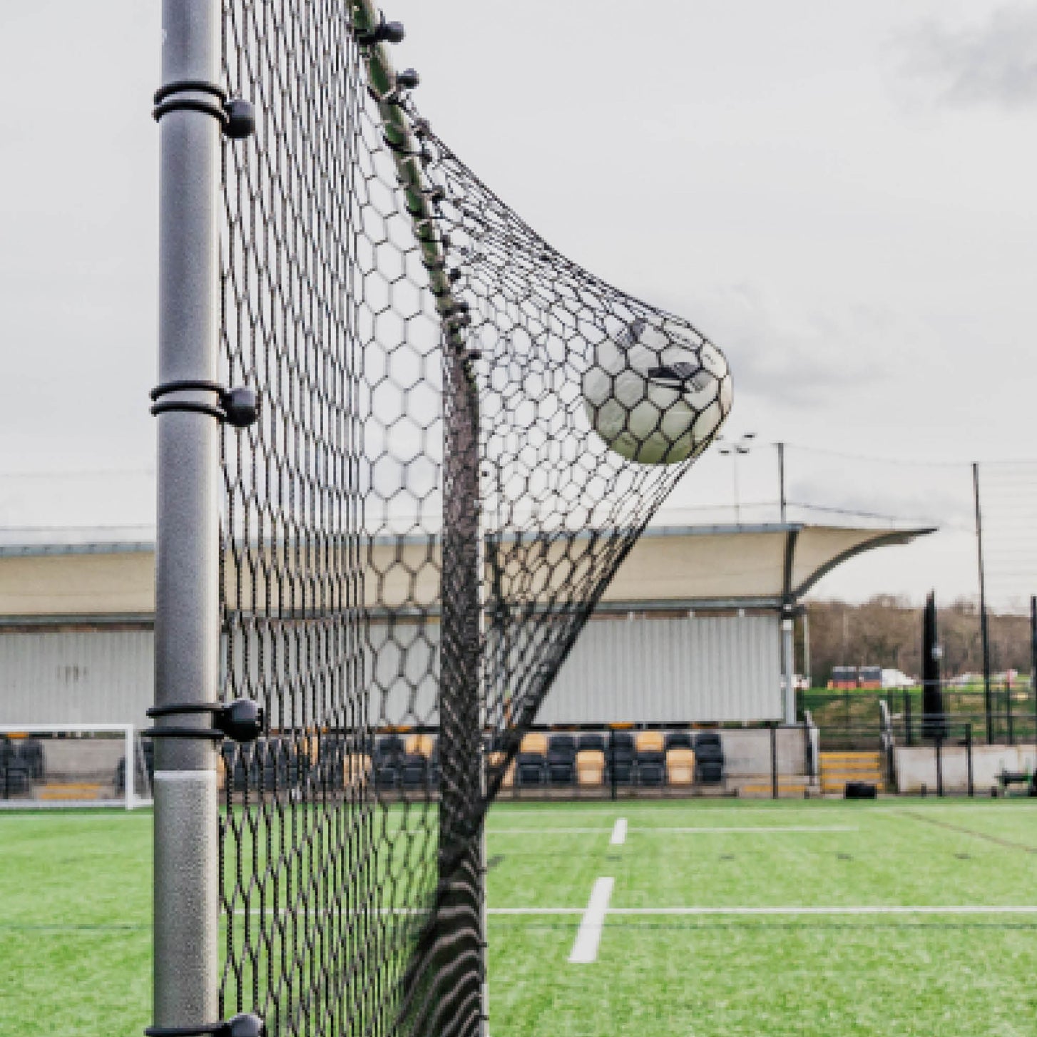 Ball stretching the black net of the Barrier Net & Rebounder 12x6' beside a grey post with net ties, seen side on.