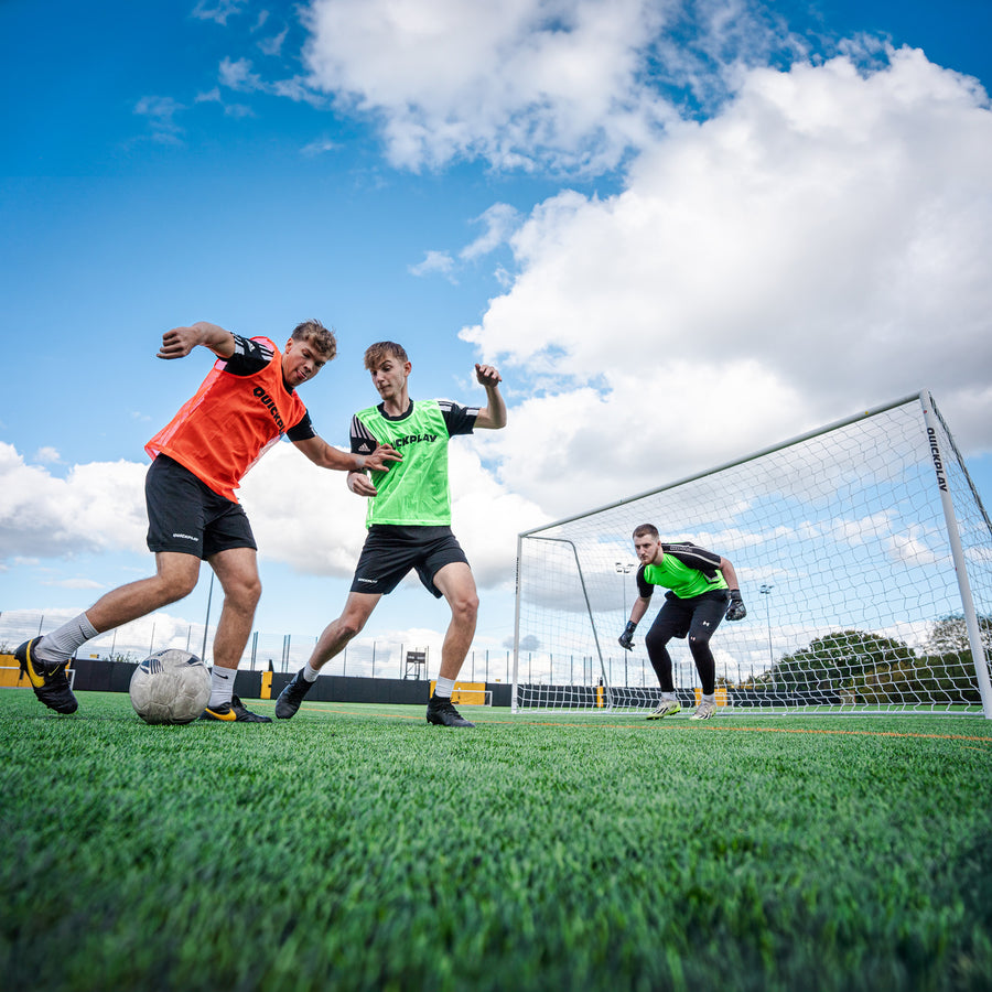 Two players in orange and green training bibs competing for the ball in front of a goalkeeper in a green bib on an artificial pitch.
