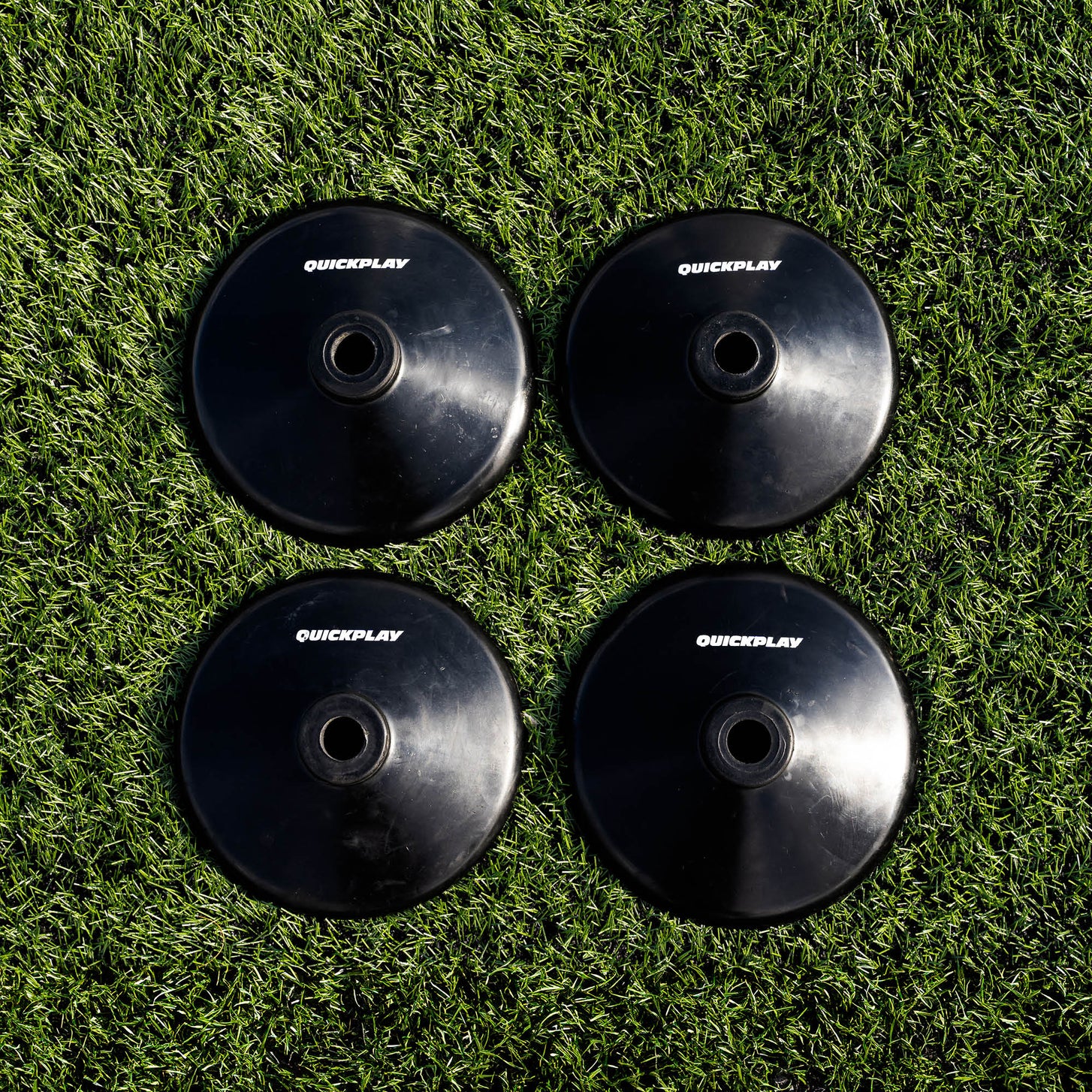 Four black round QUICKPLAY training pole rubber bases laid out in a square on artificial grass, seen from above.