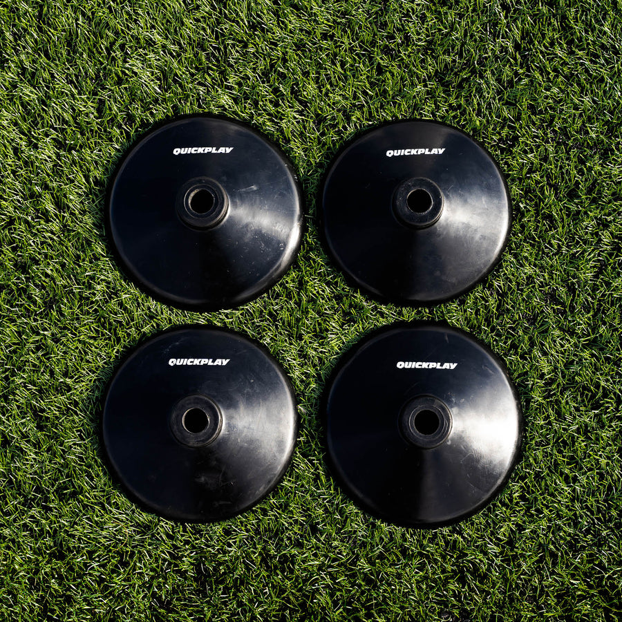 Four black round QUICKPLAY training pole rubber bases laid out in a square on artificial grass, seen from above.