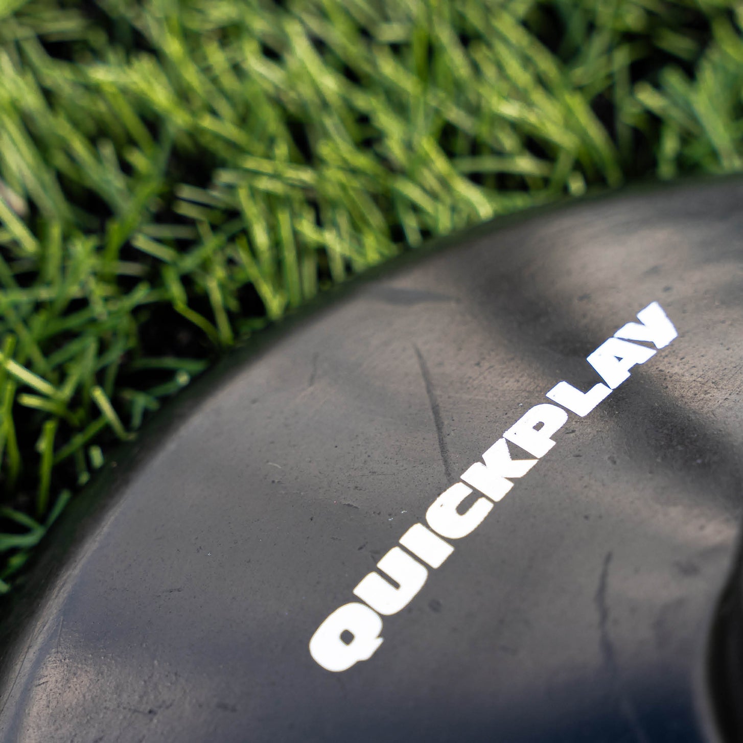 Training pole rubber base close up of the white QUICKPLAY logo on the black rubber, beside artificial grass.
