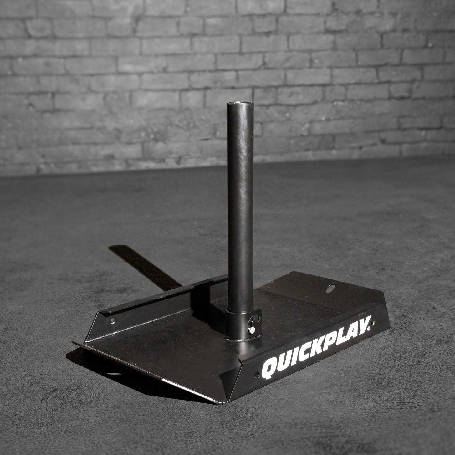 Empty black QUICKPLAY Pull Speed Sled with a central upright post, on a concrete floor against a brick wall.