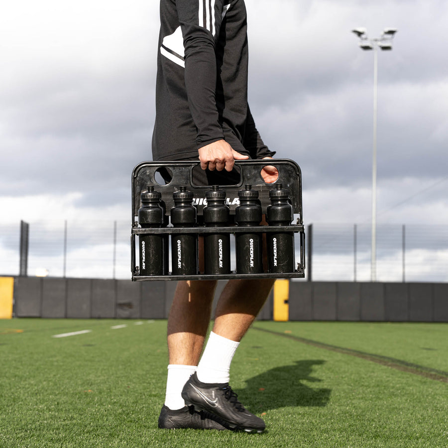 Person carrying a black crate with bottles on a sports field