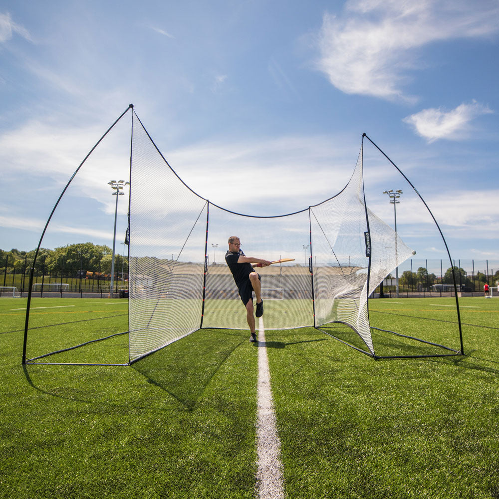 Man swinging an orange stick in front of the MEGA NET XL Ball-Stop 32x9' on an artificial pitch.