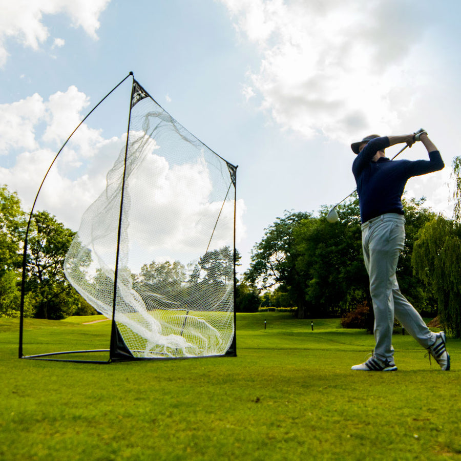Golfer swinging a driver beside the QUICK-HIT Golf Hitting Net 8x8' on a golf course, with balls in the net.