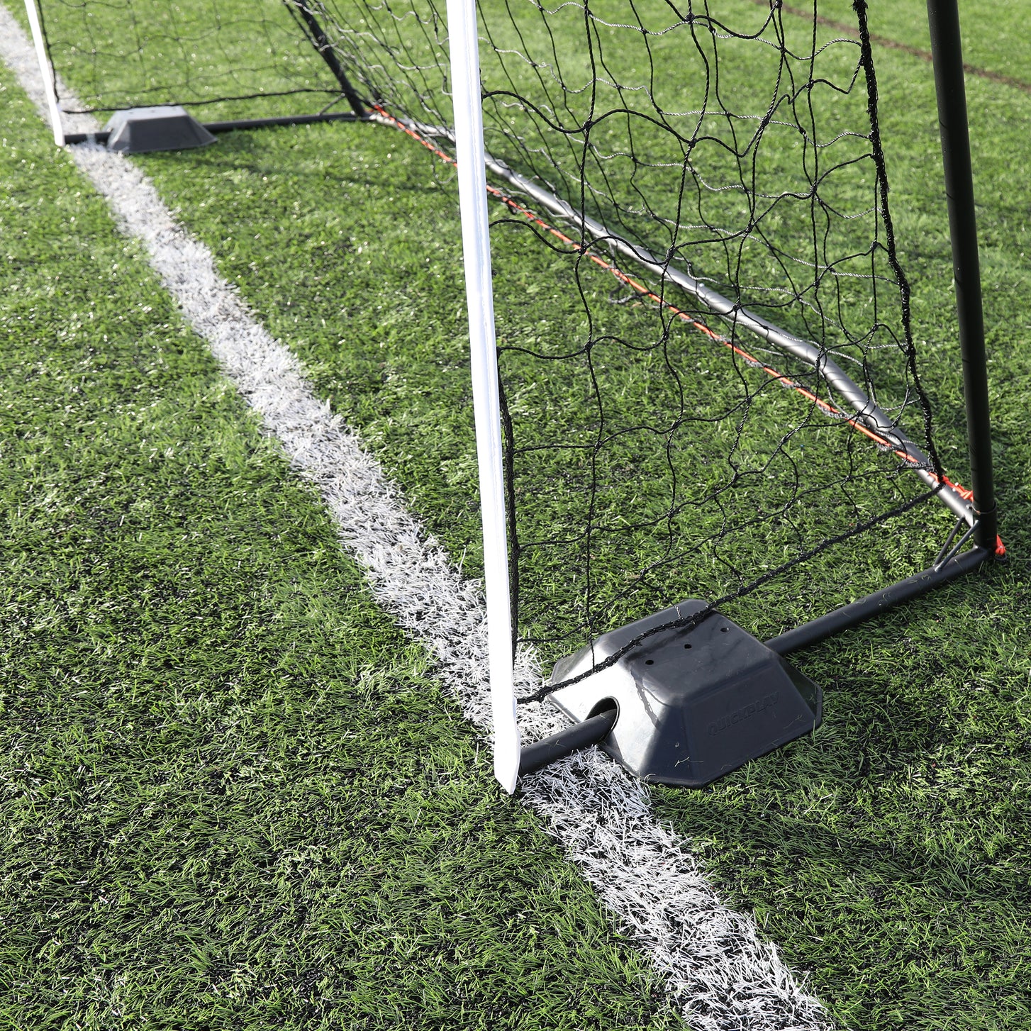 Black QUICKPLAY base weight holding the frame of a goal with a white post on artificial grass, beside a white line.