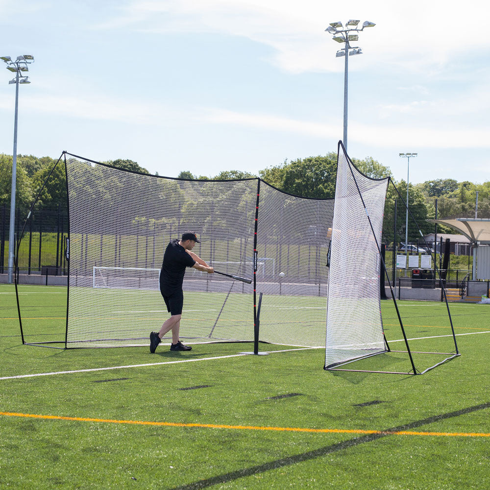Man hitting a ball with a bat into the MEGA NET XL Ball-Stop 32x9' on an artificial pitch, seen from the side.