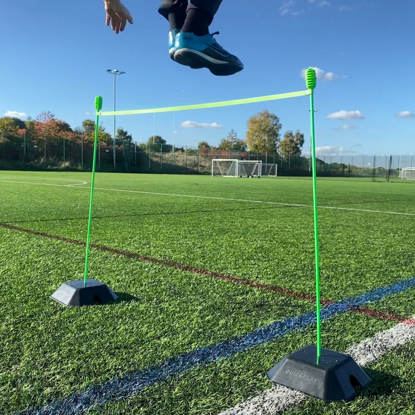 Child jumping over a green hurdle bar held by two green poles set in black QUICKPLAY base weights on artificial grass.