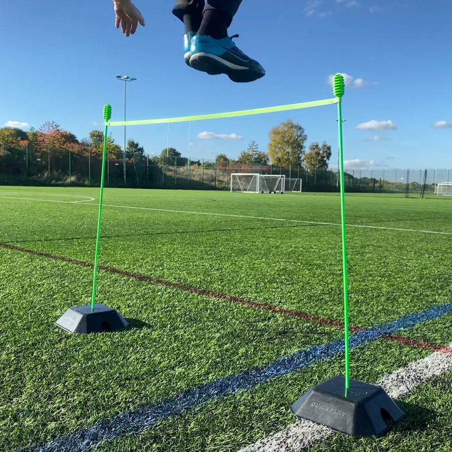 Child jumping over a green hurdle bar held by two green poles set in black QUICKPLAY base weights on artificial grass.