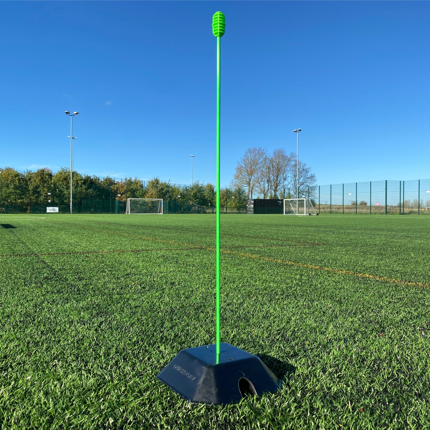 Single tall green pole standing in a black QUICKPLAY base weight on an artificial pitch, low angle.